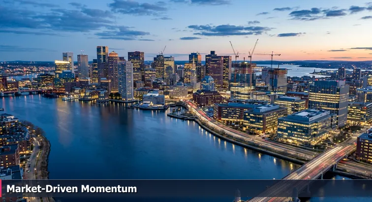 Aerial evening view of Boston’s Seaport and Kendall Square with lit office towers, construction cranes, and the Charles River in the foreground.